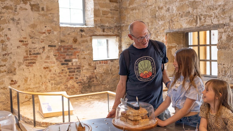 An adult white male and two white female children are standing inside a stone building with sunlight streaming through the windows. They are at a counter, on which is a tray of what look like flapjacks, which is covered with a glass dome.